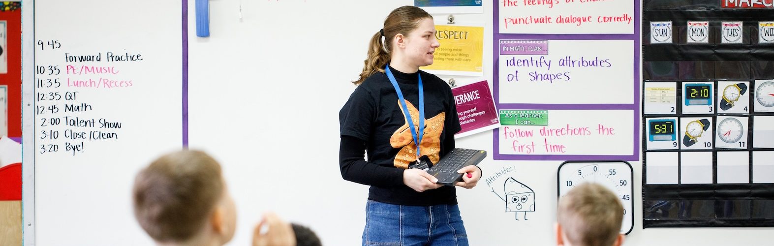 A teacher stands in front of classroom, in front of whiteboard. Students are in the foreground, with their backs to the camera.